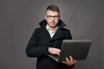business man standing in classic jacket and glass holding laptop looking at camera against gray studio background.