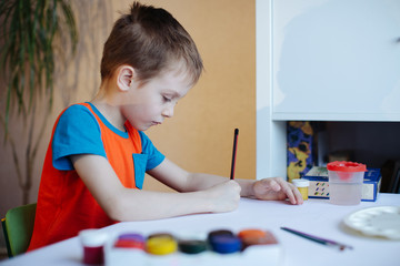 The boy draws with pencils and paints on a white sheet. near a lot of colored jars with paint. Gouache, watercolor theme.