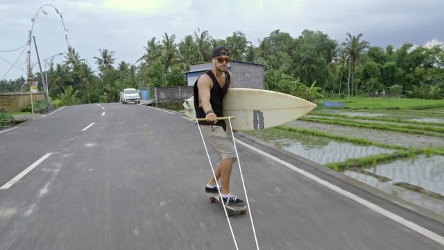 POV of young skateboarder in sunglasses skitching with surfboard along tropical road in Bali