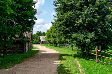 Fototapeta na wymiar Old village street in sunny summer day with ground pathway, green grass and trees, and traditional pole fence