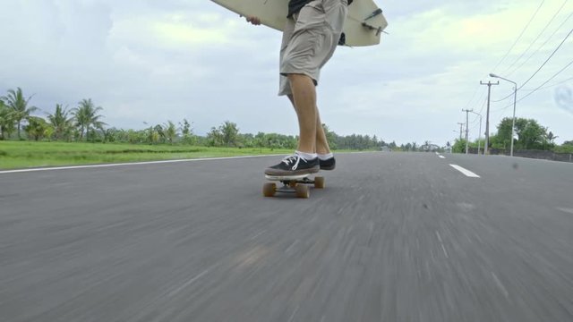 Low angle view of legs of skateboarder skitching after scooter along tropical road and holding surfboard
