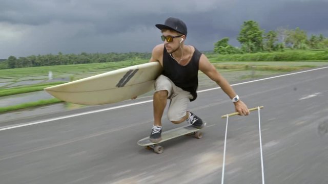 Young longboarder listening music with headphones and holding surfboard while skitching after scooter along tropical road in Bali