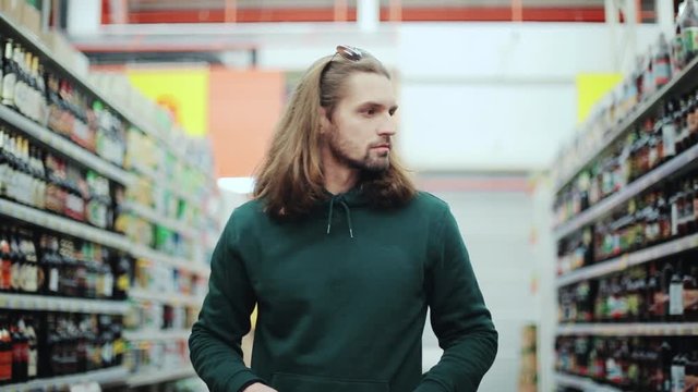 attractive man with long hair choosing bottle alcohol in the supermarket buy selection alcoholic sale handsome reading market people hand grocery looking male drink holding beer portrait slow motion