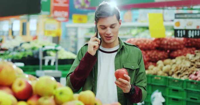 Smiling man buying an apple speak on phone in a supermarket shopping fruit sun choosing store supermarket, customer male market looking shop fresh retail adult healthy lifestyle sale young slow motion