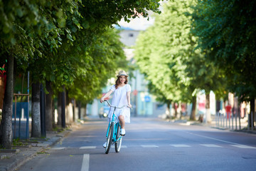 Fototapeta na wymiar Young woman rides on the road on a retro bike on a summer day. The girl is dressed in a light dress and hat on her head against a background of green urban plantings