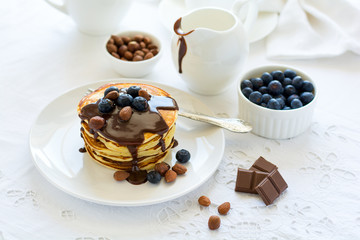Traditional breakfast concept. Stack of pancakes with chocolate sauce, blueberries and nuts on white table cloth. Selective focus