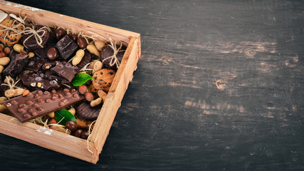 A set of milk chocolate and black chocolate in a wooden box with nuts and biscuits. On a black wooden background. Copy space for text.
