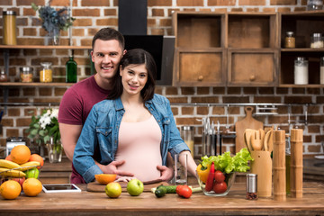 beautiful happy young pregnant couple hugging and smiling at camera in kitchen