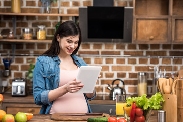 smiling young pregnant woman using digital tablet while cooking at kitchen