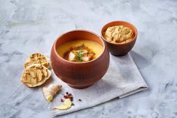 Clay pot of pumpkin soup on napkin over white textured background, close-up, selective focus, top view.