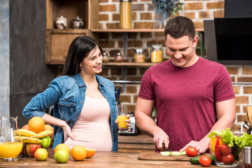 smiling young pregnant woman holding glass of fresh juice and looking at husband cutting celery at kitchen