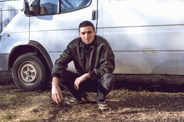 A young guy of criminal appearance in a black leather jacket stands near an old white van