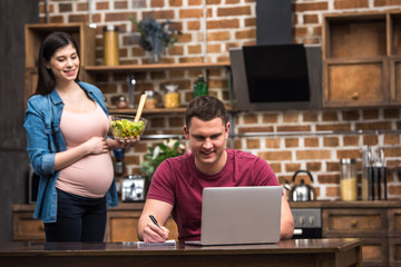 smiling young man using laptop and taking notes while pregnant wife holding glass bowl with vegetable salad