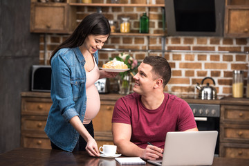 smiling young pregnant woman giving coffee and pastry to happy husband working with laptop at home