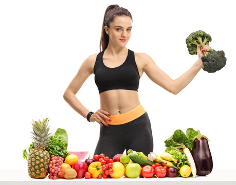 Fitness woman with a broccoli dumbbell behind a table with fruit and vegetables