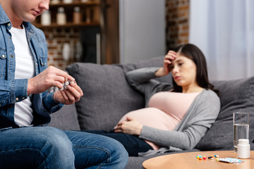 Fototapeta na wymiar cropped shot of man holding pills while sick pregnant woman lying on sofa