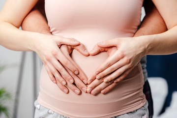 cropped shot of young pregnant couple touching belly and showing hand heart symbol