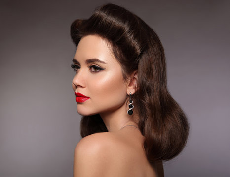Retro woman portrait. Elegant brunette with red lips makeup and pinup curly hairstyle present diamond earring jewelry isolated on gray studio background.