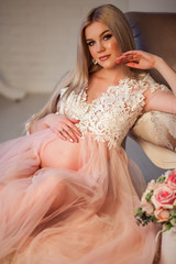 Pretty pregnant young woman is wearing luxury pink lingerie dress at home in cozy interior with bouquet of flowers