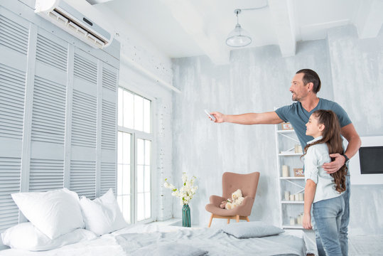 Air conditioning. Positive delighted joyful man standing together with his daughter and looking at the air conditioner while holing a remote control