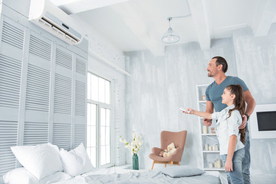 Climate control. Joyful positive nice girl standing together with her father and holding a remote control while turning on the air conditioner
