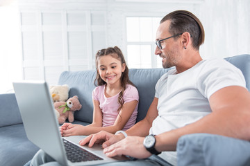 Look here. Happy delighted smart man holding a laptop and showing it to his daughter while being in a great mood