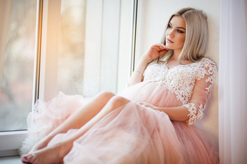 Fototapeta na wymiar Beautiful pregnant young woman is wearing luxury pink lingerie dress at home sitting on window sill, spring time