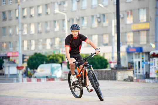 Young guy bicyclist riding on bike in cycling clothes in front of modern city building. Sportsman exercising outdoors, rest after working day. Concept of healthy life