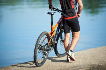 Fototapeta premium Back crop view of guy bicyclist wearing cycling clothes walking next to bicycle down pier to city lake. Sportsman resting and relaxing after training and exercising outdoors. Healthy lifestyle, nature