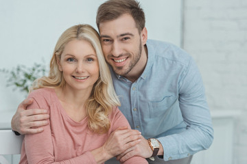 portrait of smiling grown son hugging mother and looking at camera at home
