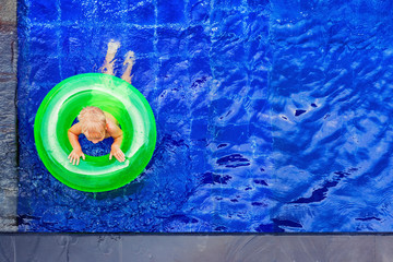 Funny photo of baby boy swimming with fun on inflatable swim ring in clear aqua park pool. Top view. Healthy family lifestyle, children with parents outdoor water sports activity on summer holidays.