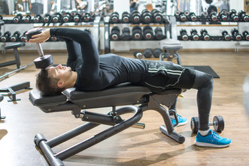 Handsome young man lying on exercise bench and lifting dumbbell in gym