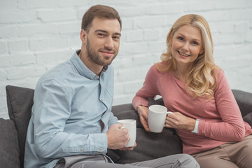 portrait of smiling mother and grown son with cups of hot drinks at home