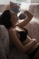 Sexy young woman in a black lingerie vaping in bedroom. Beautiful brunette woman smoking