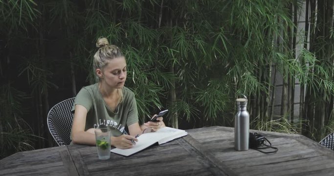 young handsome blond girl or woman (student, artist, writer) sitting and working at the table in the park, surfing internet, checking email, holding phone in her hands, downshifting