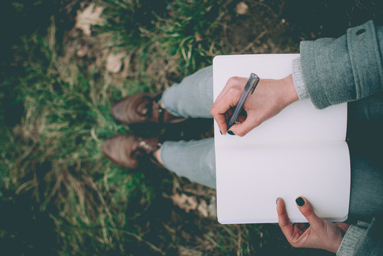 Redhead girl sitting on green spring grass writing down her thoughts in notebook. Pale hands and blank paper of notebook. Concept (idea) of thinking, dreaming, motivation, planning, studying.
