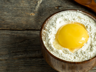Wooden bowl with flour and broken chicken egg on rustic background