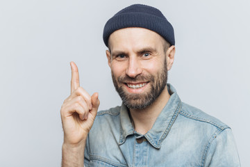 Naklejka premium Attractive middle aged male raises fore finger as gets good idea in mind, wears fashionable clothing, has pleasant smile. Businessman keeps finger up, happy to remember new successful investing plan