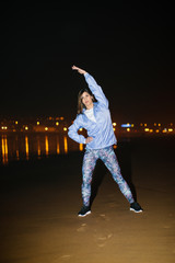 Fototapeta na wymiar Sporty young woman doing stretching exercises for warming up before fitness workout at night on a city beach. Gijon, Asturias, Spain.