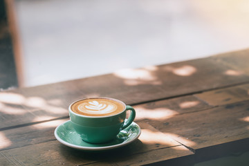 Hot cappuccino coffee in a green cup with latte art on the wooden table in a coffee shop and glass background. Vintage toned.
