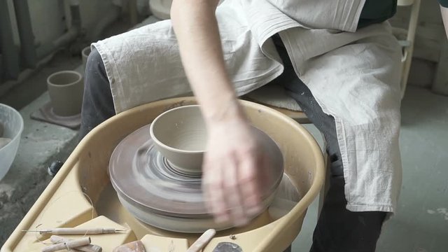 Using damp sponge potter shapes bowl on the potter's wheel. Craftsman in an apron on special equipment in workshop with his hands and tools makes a groove in the dishes of clay.