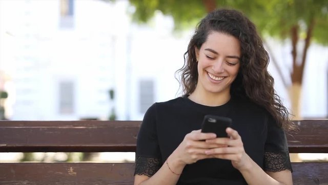 Beautiful young Caucasian woman with curly hair sending text to a friend on her smartphone and smiling