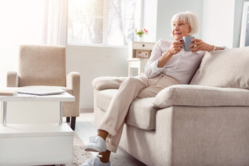 Relaxing in morning. Beautiful elderly woman sitting on the couch in her living room, drinking coffee and contemplating something