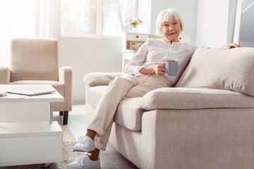 Relaxing pastime. Petite senior woman sitting on the couch, leaning comfortably against the backrest, and posing for the camera while holding a cup of coffee