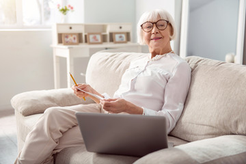 Brainstorming ideas. Charming elderly woman sitting on the sofa and posing for the camera while developing a project plan and writing down the draft on a sheet of paper