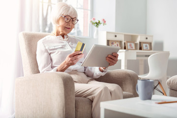 Easy and convenient. Adorable elderly woman sitting in the comfortable armchair and doing online shopping, ready to pay with her credit card