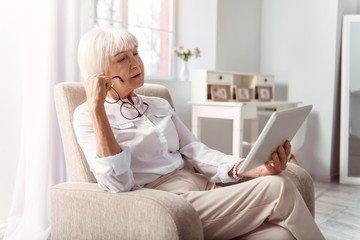 Thoughtful reader. Pleasant senior lady reading from a tablet thoughtfully, having removed her glasses, while sitting in the armchair