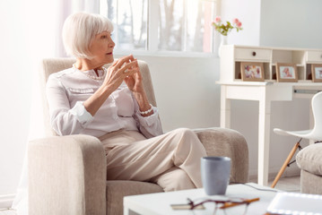 Pensive mood. Pretty elderly lady sitting in the armchair and thinking about something while pressing the fingertips on both hands together