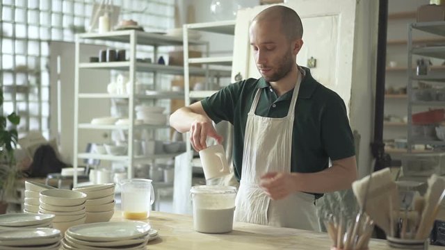 Young master immerses mug in liquid at desk pottery workshop. Man is standing at table and puts cup in container with water indoors. Bearded guy is in working process in light room with shelves on