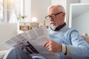 Fototapeta na wymiar Informative article. Charming elderly man in eyeglasses reading an article in the newspaper and marking interesting passages with the pencil